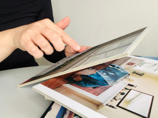 Close-up photo of a person’s hand pointing at an image in a photo album. The image shown is the living room scene from Fig03. The album is placed on a table, with multiple printed photos stacked beneath. Context suggests participatory or narrative research using visual materials.